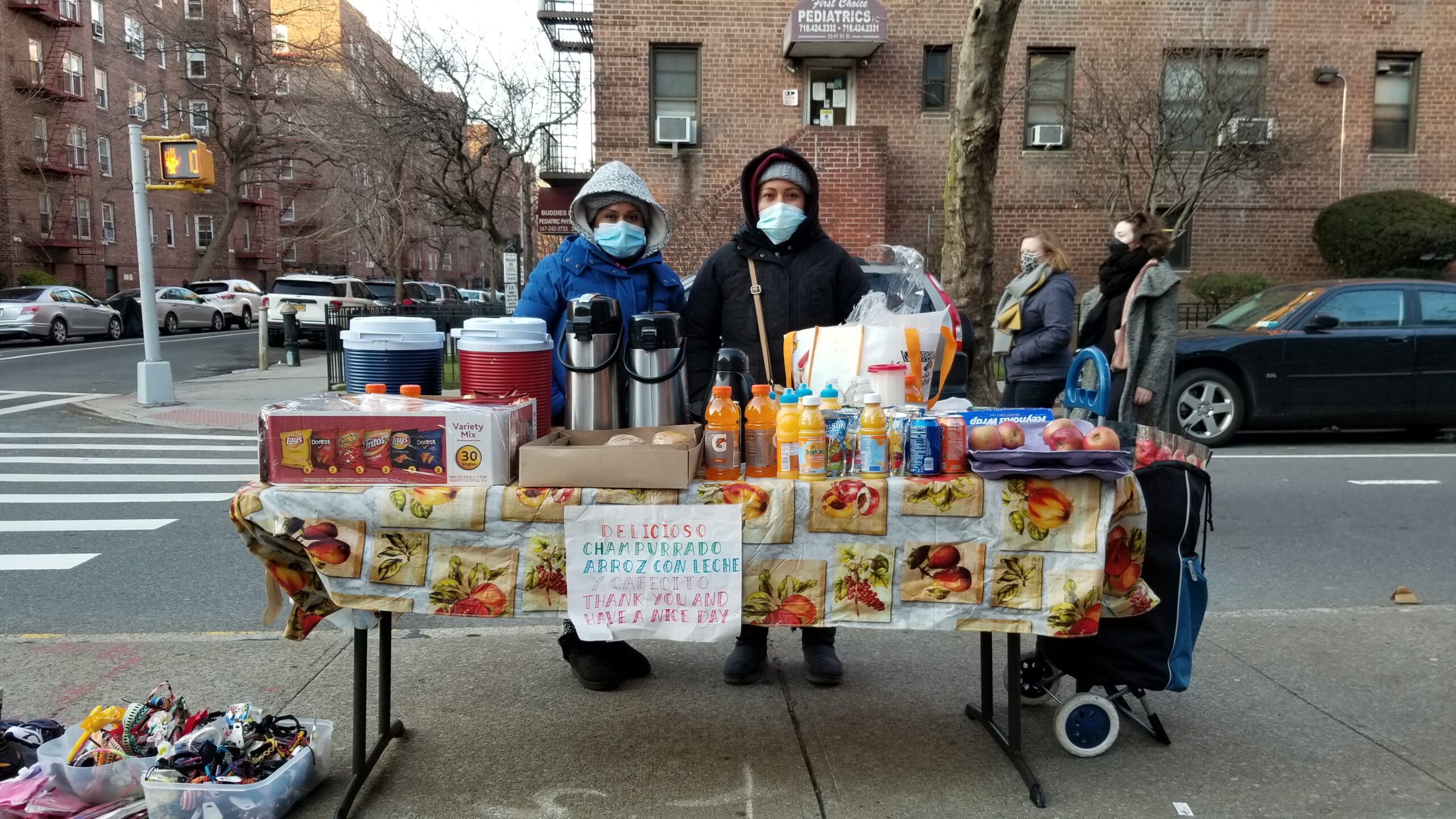 Janet Bravo stands behind her food table with her friend Hilaria. There are thermoses, drink bottles, chips, and fruit for sale. A sign read, "Delicioso Champurrado, Arroz con Leche y Cafecito. Thank you and have a nice day."