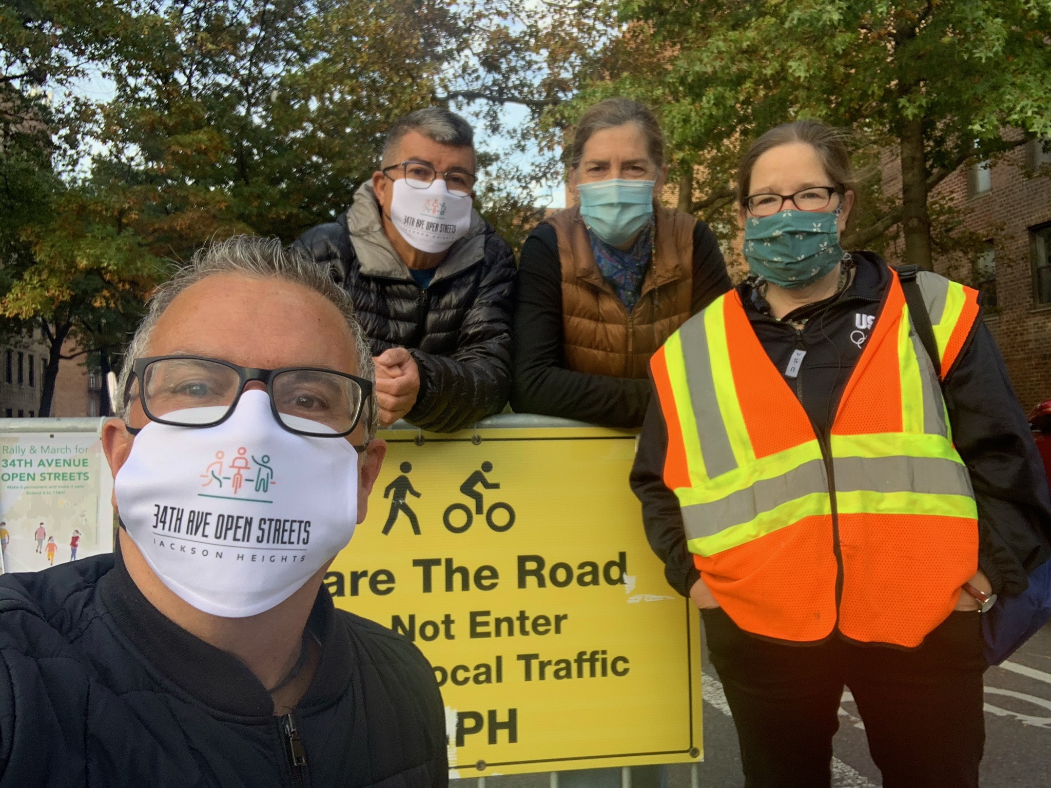 Jim Burke is taking a selfie with three friends leaning on a barricade on 34th Avenue. Two wear masks that say "34th Ave Open Streets." One is wearing a fluorescent safety vest.