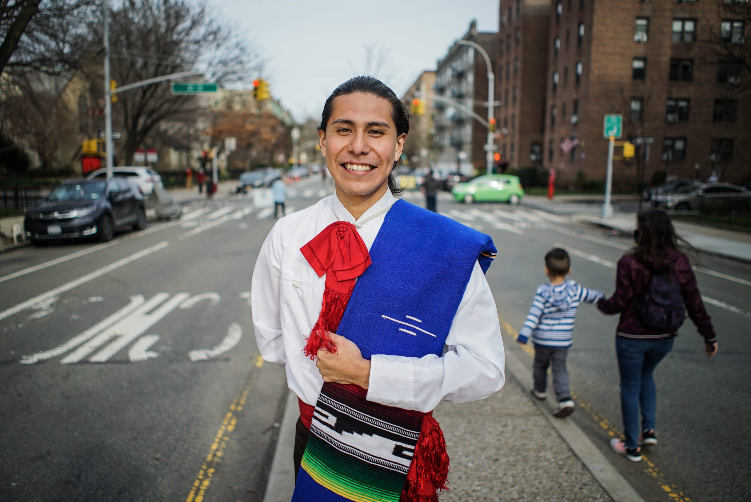 Erick Modesto stands on the median of 34th Avenue smiling and looking into the camera. Behind him, a boy balances on the curb of the median while a woman holds his hand. Erick is wearing a traditional ballet folklorico costume.