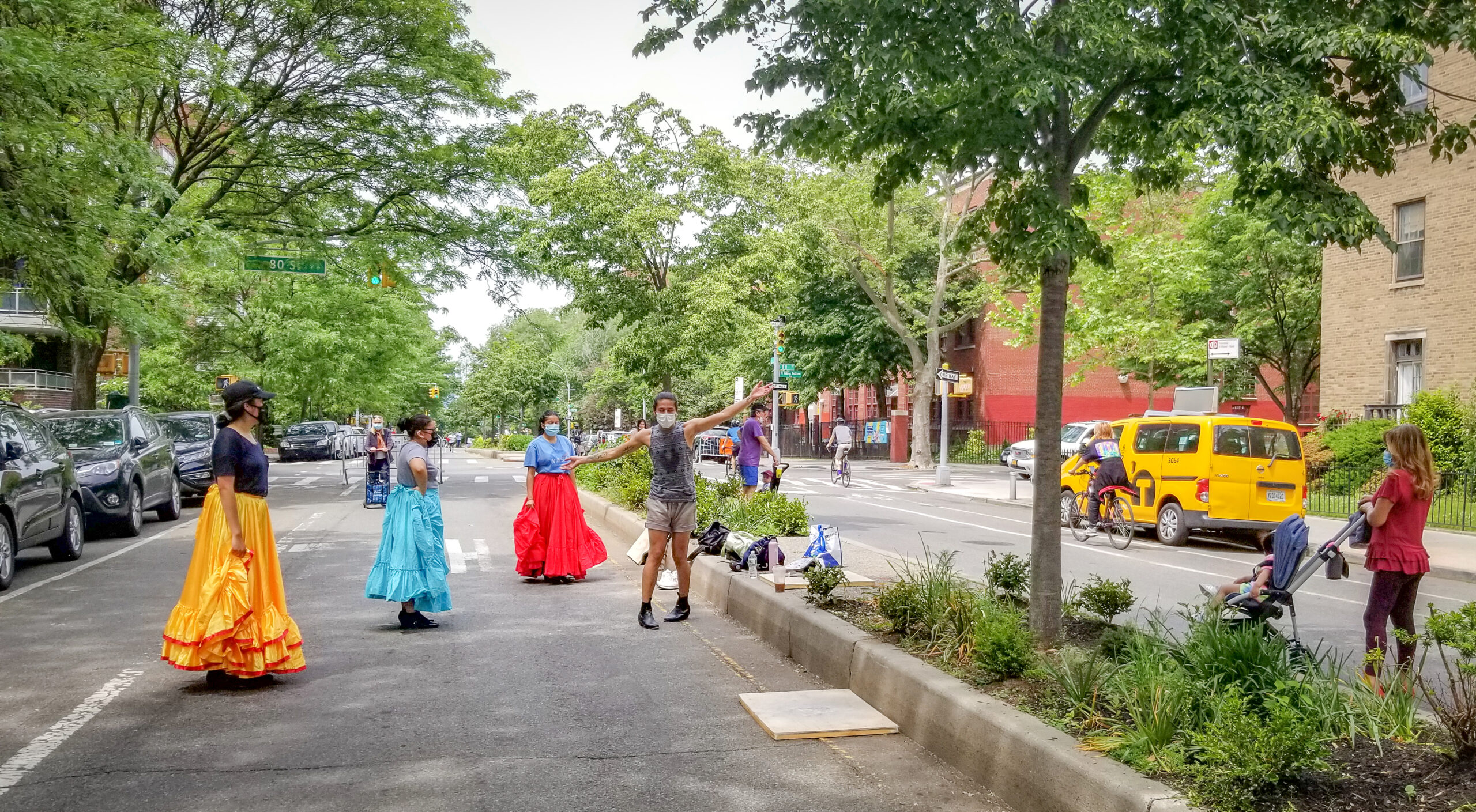 During a rehearsal on 34th Avenue, Erick demonstrates a dance move as three dancers watch him. On the other side of the median, a woman has stopped to observe the rehearsal.