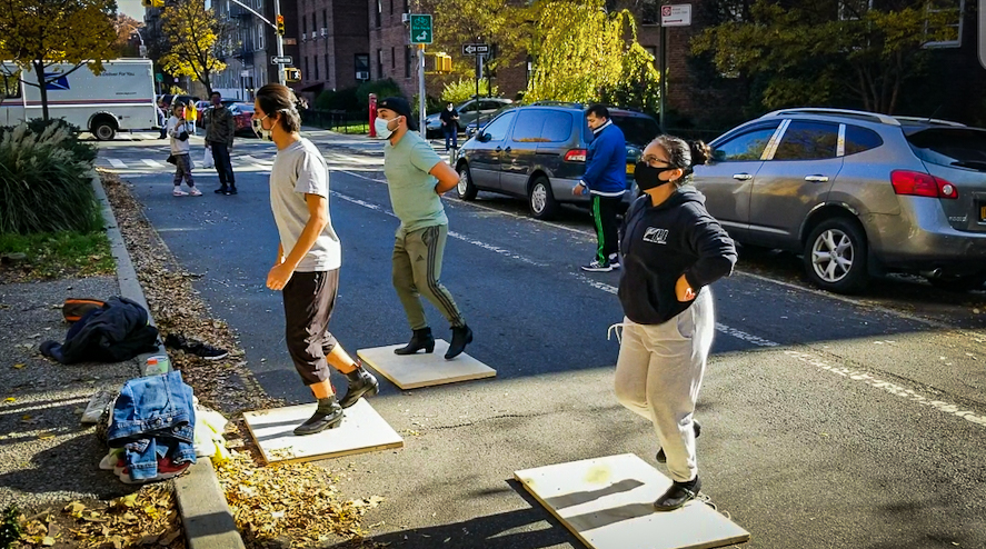 Erick and two dance troupe members rehearse on 34th Avenue. They all stand on wooden boards. It is an autumn day and there are fallen leaves on the median.