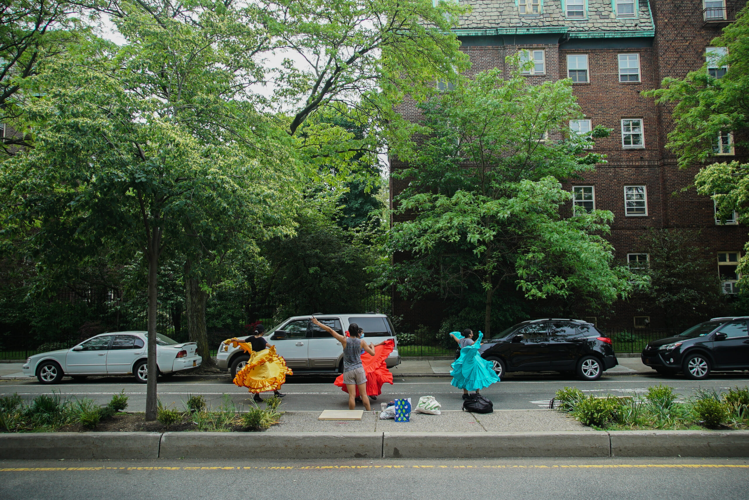 Erick and three dancers are in the middle of a move as they rehearse on 34th Avenue. The dancers are spinning and their colorful skirts are swirling around them. Erick stands in front with outstretched arms, directing them. Behind them are tall trees and apartment buildings.