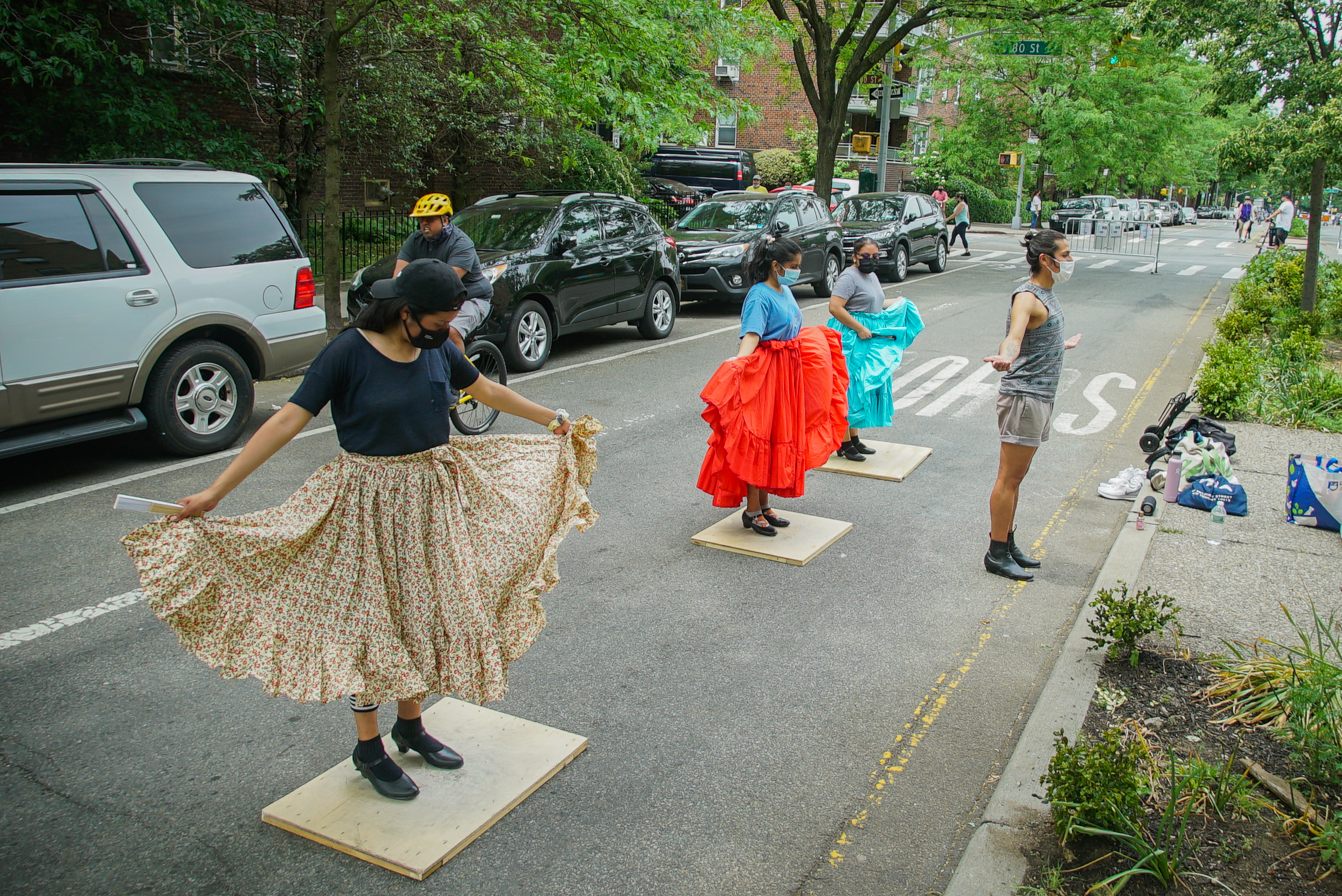 Rehearsing on 34th Avenue, three dancers stand on small wooden platforms and hold their skirts, preparing to practice steps in their routine. Erick stands in front of them and demonstrates the pose.