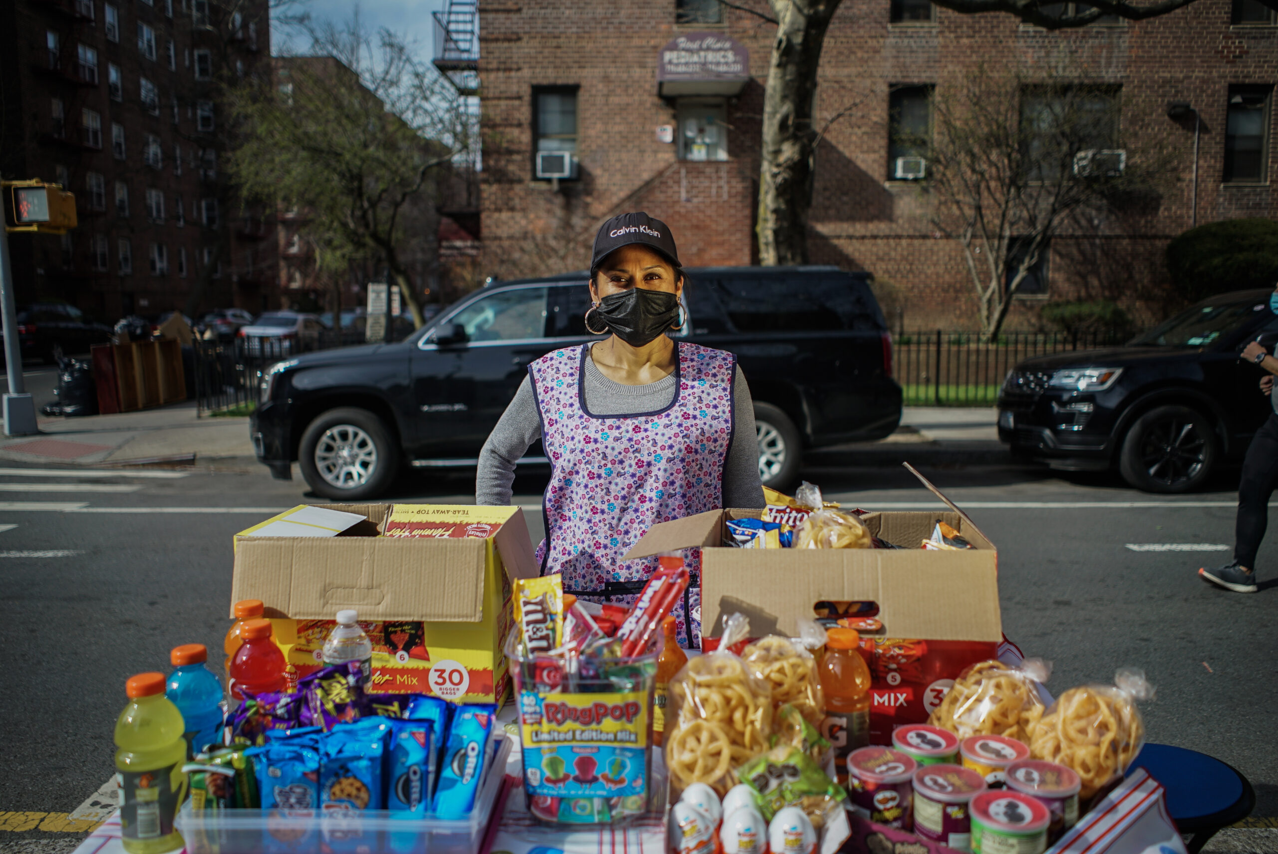 Janet Bravo stands behind her food table, wearing a mask and apron. She is selling drinks, snacks and candy.