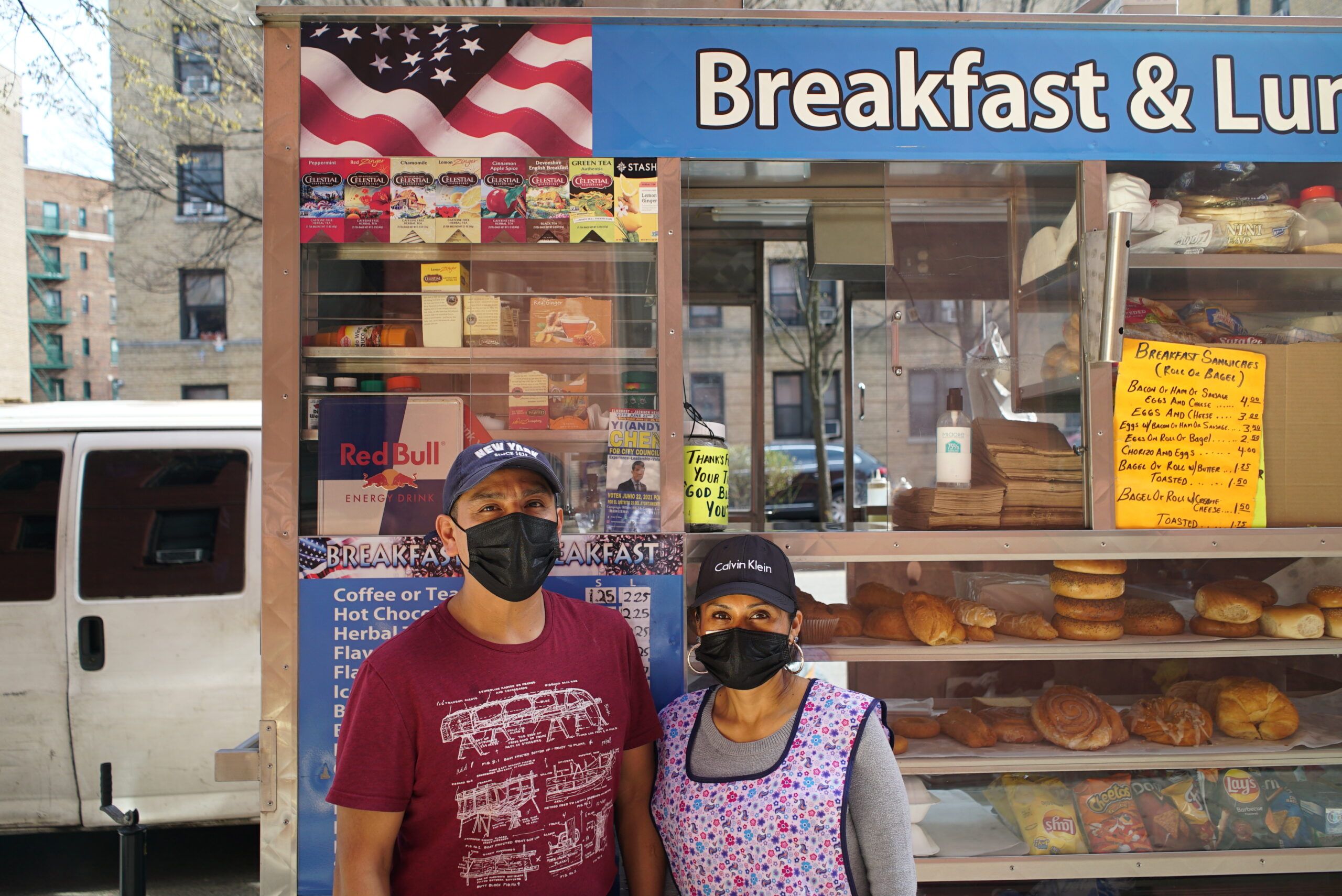 Janet and her husband Miguel are wearing masks and standing in front of their food truck, looking into the camera. Behind the glass display of the food truck there are boxes of tea, danishes, bagels, and chips. There is a menu offering different kinds of breakfast sandwiches.