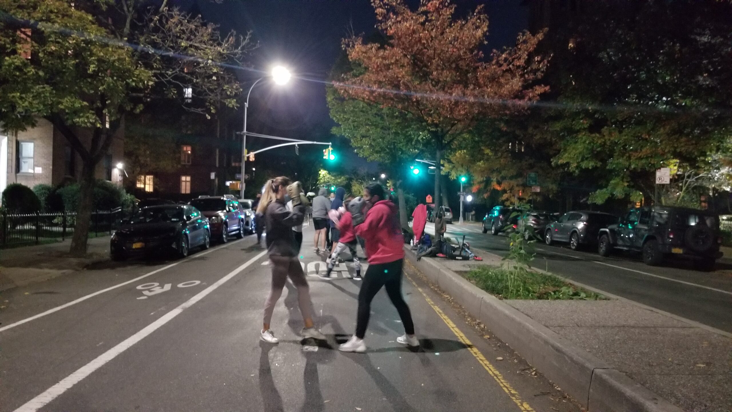 On a darkened 34th Avenue lit by streetlamps, students wearing boxing gloves are practicing kickboxing routines in the middle of the street. There are trees with autumn leaves on the median behind them.