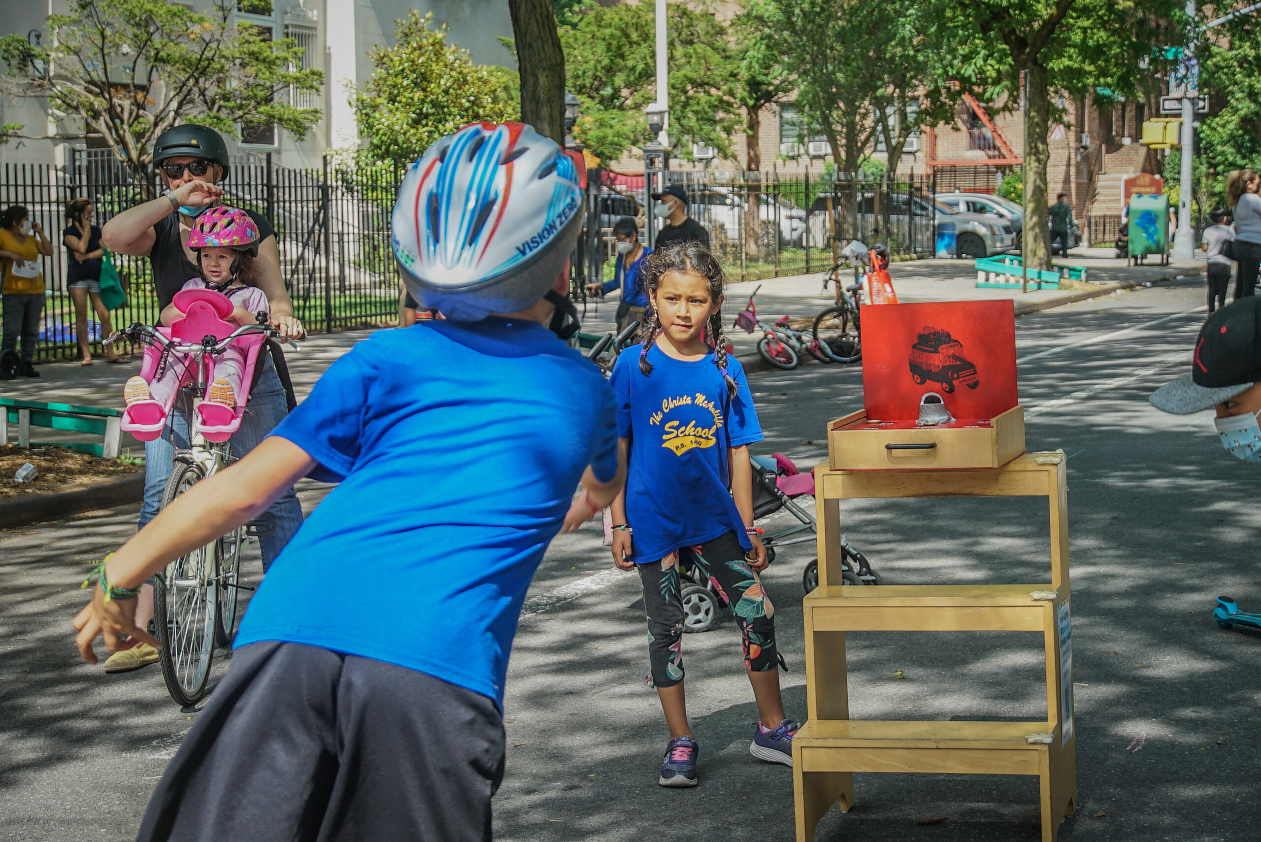 A young boy wearing a blue shirt and a bicycle helmet is throwing a coin towards the sapo board, which resembles a small suitcase with the top open, and inside there is a metal frog with an open mouth. There are many people on 34th Avenue. A young girl watches the sapo game. A mother rides by on a bike with a child strapped in.