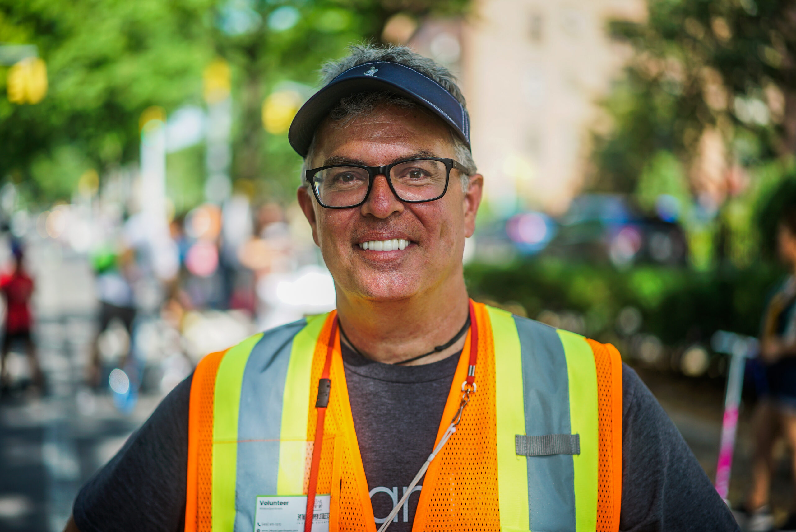 Jim Burke stands on 34th Avenue, looking at the camera and smiling. He wears a visor and a bright orange, high visibility safety vest. The sun is shining and figures of people are behind him.