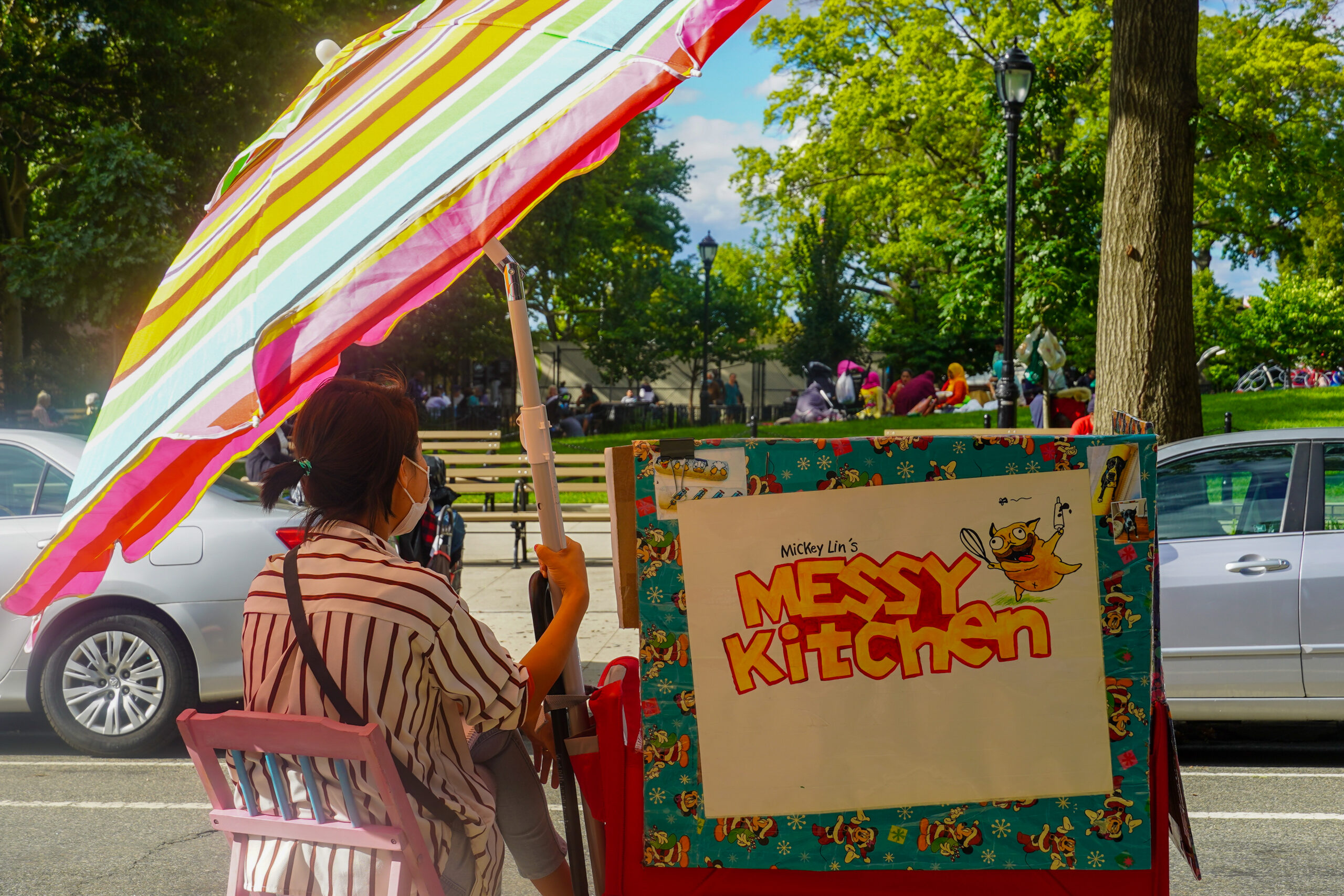 Mickey sits at her station in front of Travers Park. It's a sunny day, and she sits beneath her umbrella. The "Mickey Lin's Messy Kitchen" logo is visible on back of the wagon. In the background, people in Travers Park are playing in the handball court, and others are sitting on the grass and having a picnic.