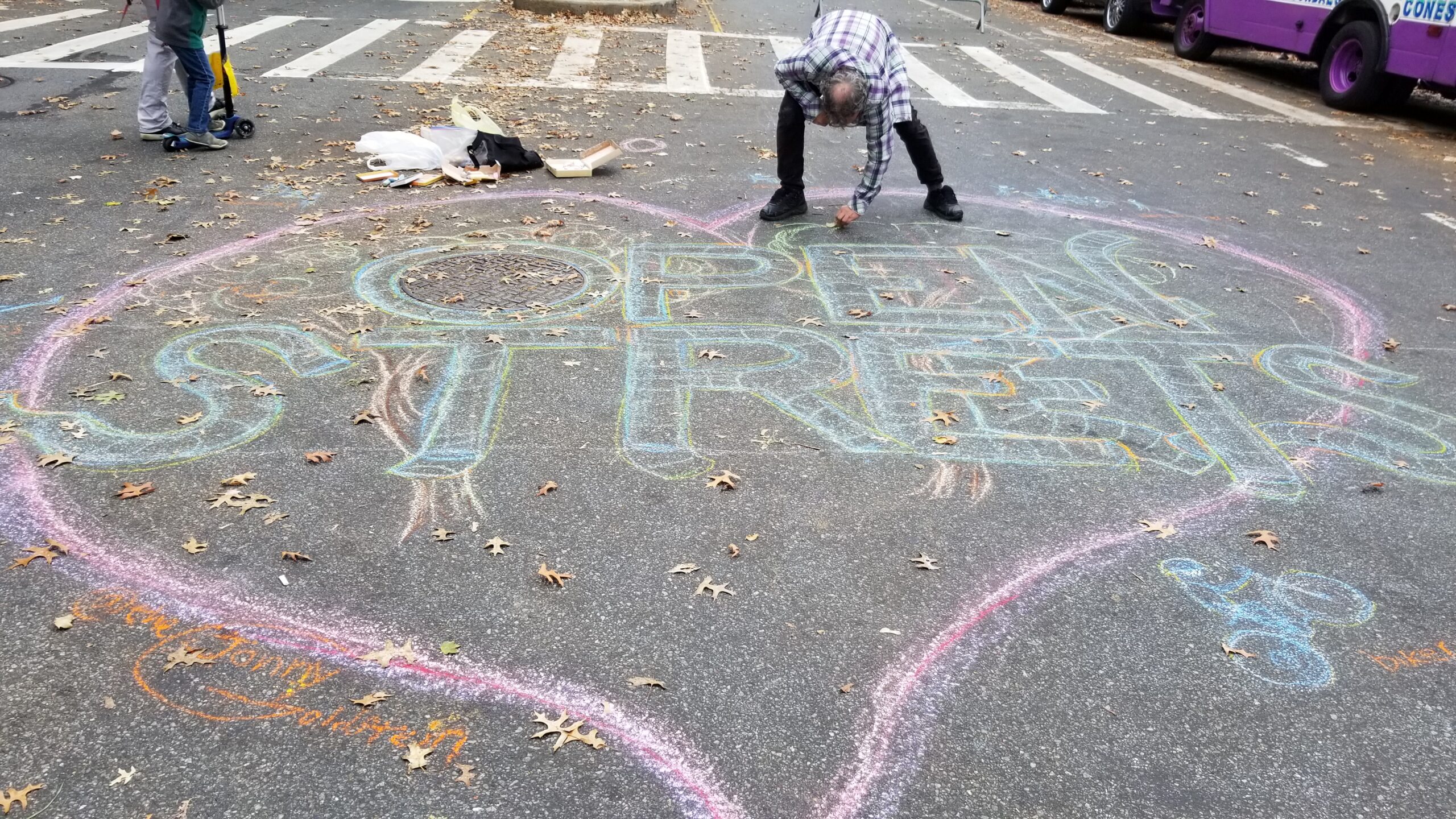 A man is bending down and writing with chalk on 34th Avenue. He is finishing a very large drawing of a heart with the words "Open Streets" inside it, and a drawing of a tree and a person on a bicylce.