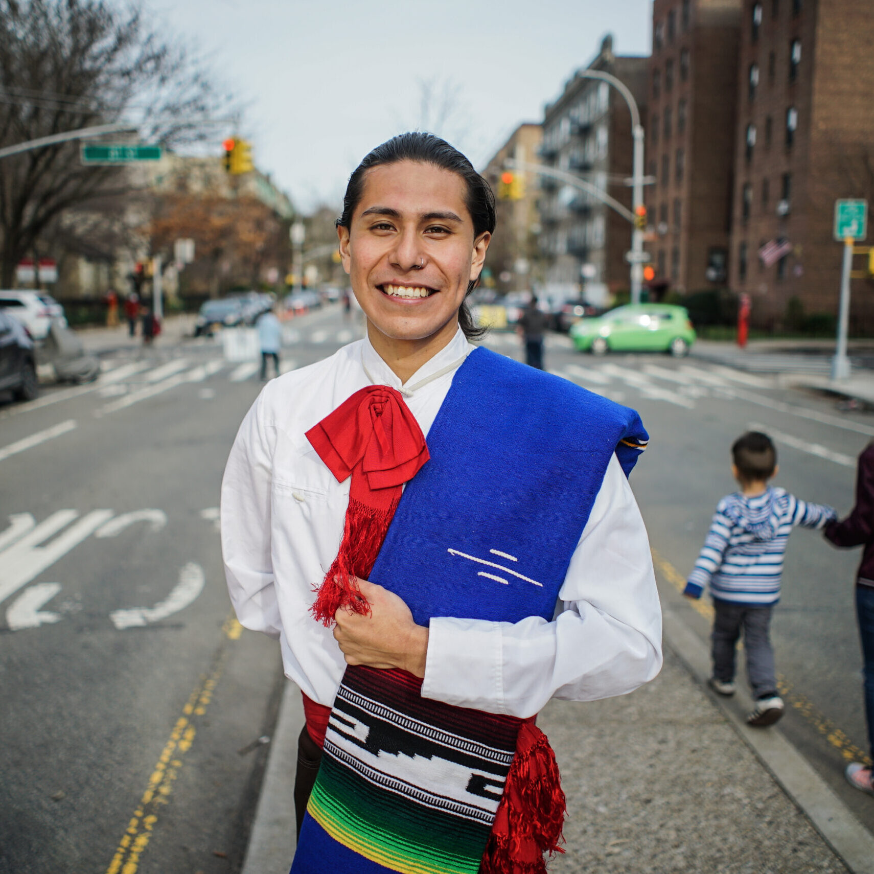 Erick Modesto stands on the median of 34th Avenue smiling and looking into the camera. Behind him, a boy balances on the curb of the median while a woman holds his hand. Erick is wearing a traditional ballet folklorico costume.