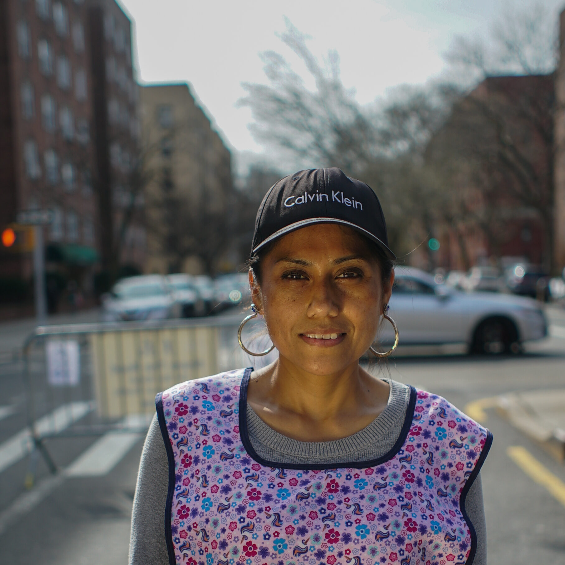 Janet Bravo stands on 34th Avenue, looking into the camera, wearing a baseball cap and apron.